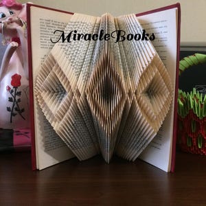 May include: A book art sculpture with a diamond pattern created by folding the pages of a book. The book is bound in red and the pages are cream coloured. The sculpture is titled "Miracle Books".