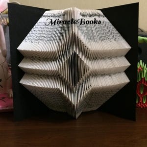 Peut inclure: Une sculpture en livre plié composée de pages blanches avec du texte noir, minutieusement pliées pour former un motif géométrique en losange. Les mots Miracle Books sont imprimés en noir sur la partie supérieure, encadrés par deux panneaux noirs.