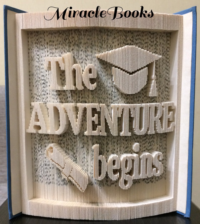 May include: A book art sculpture with the words "The Adventure Begins" in a 3D design. The sculpture also includes a graduation cap and a scroll.