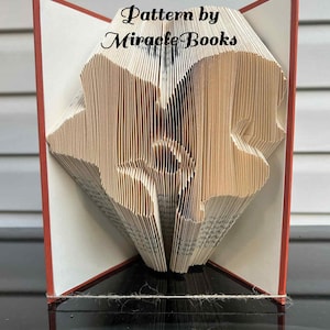 Könnte beinhalten: Ein gefaltetes Buchkunstwerk mit einem herzförmigen Ausschnitt in den Seiten. Das Buch hat rote Einbände und der Text "Pattern by Miracle Books" ist sichtbar. Die Seiten sind beige und das Buch steht auf einer reflektierenden Oberfläche.
