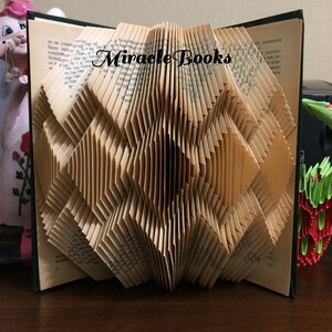 Op de afbeelding: Een open boek met gevouwen pagina's die een geometrisch patroon vormen. De pagina's zijn bruin en de kaft is donkergroen. De tekst "Miracle Books" is zichtbaar op de pagina's. Het boek staat op een houten oppervlak.