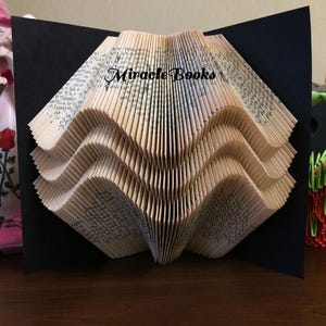 Puede incluir: Escultura de libro plegado en forma de olas. El libro abierto muestra el título "Miracle Books". La escultura se presenta sobre un fondo negro, creando un contraste llamativo.