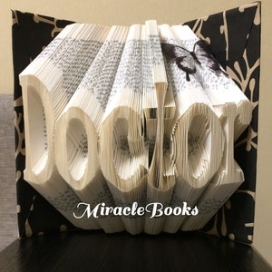 May include: A folded book art sculpture with pages shaped to spell the word "doctor" in a serif font. The book features a black and white butterfly graphic on the top right page and is framed by black covers with a white branch pattern.