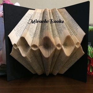 May include: A unique book art piece featuring folded pages in a diamond pattern, set against a black background. The text "Miracle Books" is visible on the pages. The artwork is displayed on a wooden surface.