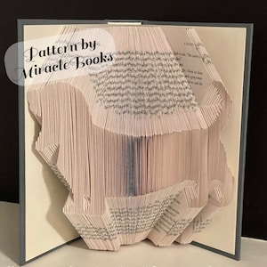 Peut inclure: Une sculpture de livre avec des pages pliées formant la silhouette d'un lion. Les pages sont de couleur crème avec du texte imprimé en noir. Une superposition semi-transparente à gauche affiche le texte Pattern by Miracle Books.