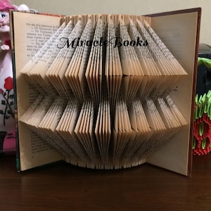 Op de afbeelding: Een boek met pagina's gevouwen tot een symmetrische, geometrische sculptuur. Het papier is crèmekleurig met zwarte gedrukte tekst. De woorden Miracle Books staan in zwarte schrijfletters gedrukt over het midden van de gevouwen pagina's.