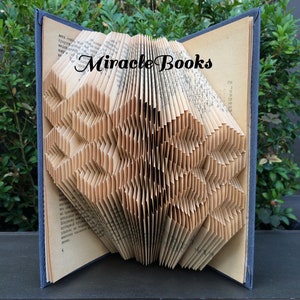 Op de afbeelding: Een boekkunst sculptuur in de vorm van een sneeuwvlok, gemaakt door de pagina's van een boek te vouwen. Het boek is open en de pagina's zijn gevouwen tot een driedimensionale vorm. Het boek ligt op een donker oppervlak. De titel "Miracle Books" is zichtbaar op het boek.