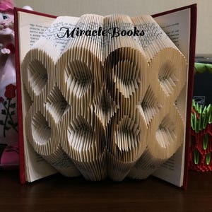 Puede incluir: Una escultura de libro plegado con cubiertas rojas, con páginas intrincadamente dobladas para formar un patrón geométrico repetitivo de formas circulares. El texto Miracle Books está impreso en negro en la parte superior central.