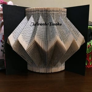 Könnte beinhalten: Eine handgefertigte Buchskulptur mit gefalteten Seiten, die ein rautenförmiges Design bilden. Die Skulptur befindet sich vor einem schwarzen Hintergrund, mit dem Text "Miracle Books" auf den Seiten. Die Buchseiten sind beige.
