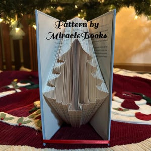 May include: A white book folded into the shape of a Christmas tree. The book is standing on a red and white patterned surface. The text on the book reads "Pattern by Miracle Books".