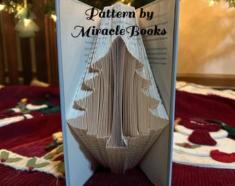 May include: A folded book art sculpture featuring a Christmas tree shape created by precisely folding the pages. The text Pattern by Miracle Books is printed across the center. The book has a light blue cover and a thin red ribbon at the base.