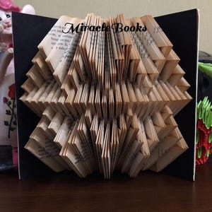 Può includere: Una scultura di libro piegato con pagine che formano un design geometrico. Il libro è aperto, rivelando le pagine piegate in un motivo simmetrico. Il testo "Miracle Books" è visibile nella parte superiore del libro.