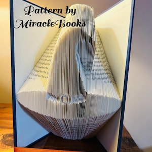 Op de afbeelding: Een boeksculptuur met gevouwen pagina's die een paard silhouet creëren. Het boek is open, met een donkerblauwe kaft en een oranje lint. De tekst "Pattern by Miracle Books" is zichtbaar.