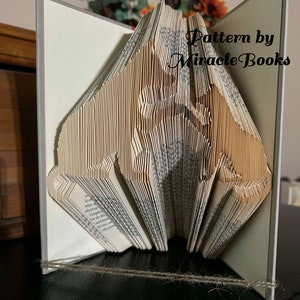 May include: A book folded into the shape of two dogs, one standing on its hind legs and the other sitting. The book is open and the spine is visible. The book is on a table with a brown surface. The text "Pattern by Miracle Books" is visible on the right side of the book.