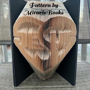 May include: A folded book art sculpture featuring the word 'Jesus' shaped into a heart. The pages are intricately creased to form the letters and heart outline. The text 'Pattern by Miracle Books' is printed in black across the top of the pages.