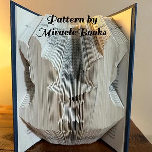 May include: A folded book art sculpture featuring a central star shape and two flanking silhouettes, with the text Pattern by Miracle Books overlaid. The book has dark blue covers and white pages that are precisely folded to create the design.