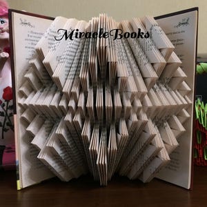 Peut inclure: Un livre ouvert dont les pages sont pliées en un motif géométrique. Les pages sont beiges, et les bords sont brun foncé. Le texte "Miracle Books" est imprimé sur les pages. Le livre est posé sur une surface en bois.