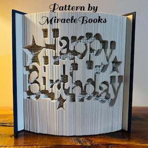 Puede incluir: Un libro con las palabras "Happy Birthday" recortadas de las páginas. El libro está doblado para crear un efecto 3D. El libro está sobre una superficie de madera. El texto "Pattern by Miracle Books" está en la parte superior de la imagen.