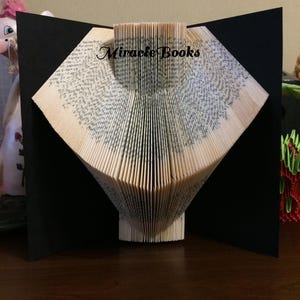 Peut inclure: Une sculpture de livre plié avec le texte "Miracle Books" sur les pages. Le livre est plié en forme de losange, avec les pages déployées. Le fond est noir et la sculpture est sur une surface marron foncé.