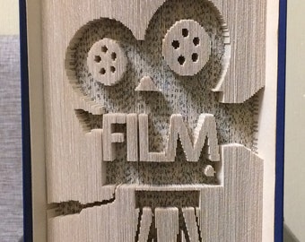 May include: A book art sculpture of a vintage film camera with the word "FILM!" in the centre. The sculpture is made from the pages of a book that have been carefully folded and cut to create the design. The book is bound in blue and has the words "Miracle Books" printed on the bottom.