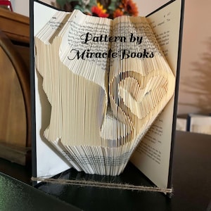 Może przedstawiać: Książka z pozaginanymi stronami tworzącymi sylwetkę kota i kształt serca. Tekst Pattern by Miracle Books jest nadrukowany na środku stron. Książka ma czarną okładkę i jest przytrzymywana przez cienki sznurek.