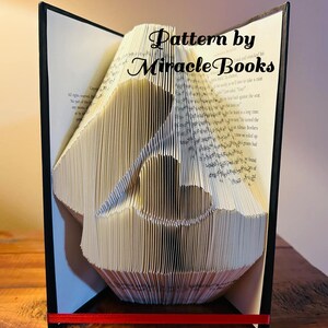 May include: A book art sculpture of a bird, folded from the pages of a book. The book is open and the sculpture is standing upright. The book is bound in black and has a red ribbon tied around it. The text "Pattern by Miracle Books" is visible on the book.