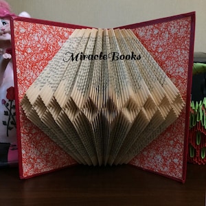 May include: An open book with pages folded into a fan shape. The book's cover is red with a white and red pattern. The text "Miracle Books" is visible on the pages. The book is displayed on a wooden surface.