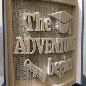 May include: A book with the words "The Adventure Begins" folded into the pages. A graduation cap and a pencil are also folded into the pages. The book is blue and has the words "Miracle Books" printed on the bottom.