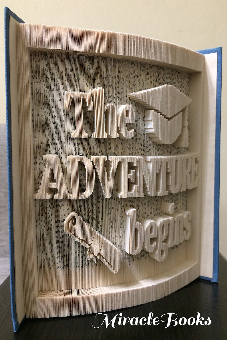 May include: A book with the words "The Adventure Begins" folded into the pages. A graduation cap and a pencil are also folded into the pages. The book is blue and has the words "Miracle Books" printed on the bottom.
