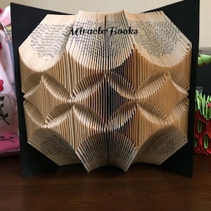 Può includere: Scultura di libro piegato con la scritta "Miracle Books" in alto. Le pagine sono piegate per creare un disegno geometrico. Il libro è ambientato su uno sfondo nero.