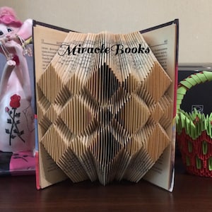 May include: A decorative book sculpture with folded pages forming a diamond pattern. The open book displays the text "Miracle Books". The pages are beige and brown, creating a textured effect. The book is a unique art piece.