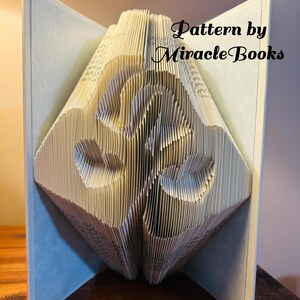 Op de afbeelding: Een gevouwen boekkunstsculptuur met twee handen die een hartvorm vormen, gemaakt door de pagina's van een boek nauwkeurig te vouwen. De tekst "Pattern by MiracleBooks" is zichtbaar in zwart schrift aan de rechterbovenkant van de witte pagina's.