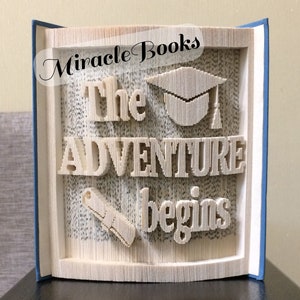 May include: A book art sculpture with the words "The Adventure Begins" and a graduation cap and diploma. The sculpture is made from the pages of a book and is framed in a blue book cover.