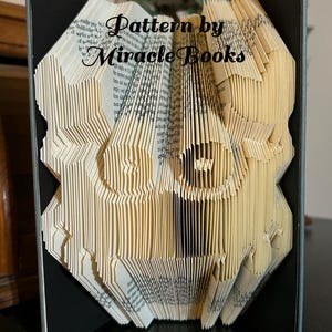 Op de afbeelding: Een gevouwen boekkunstwerk met een geometrisch ontwerp, weergegeven in een zwart frame. De pagina's zijn beige en de tekst "Pattern by Miracle Books" is zichtbaar. Het kunstwerk staat tegen een neutrale achtergrond.