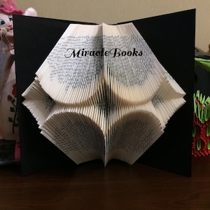 May include: A folded book sculpture with the text "Miracle Books" on the pages. The pages are folded to create a three-dimensional design against a black backdrop. The book is open, revealing the intricate folds.