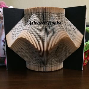 Puede incluir: Escultura de libro hecha a mano con páginas plegadas en forma de abanico. El texto "Miracle Books" está impreso en las páginas. La escultura se exhibe sobre un fondo negro.