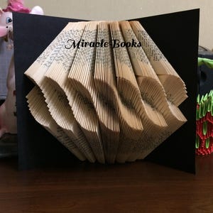 May include: A handmade book sculpture with pages folded into a wave pattern. The book is set against a black background, with the text "Miracle Books" visible on the pages. The sculpture is a unique decorative item.