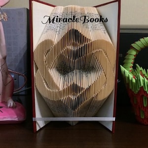 Könnte beinhalten: Eine gefaltete Buchkunst-Skulptur, deren Seiten zu einer Herzform geformt sind. Das Buch hat einen roten Einband und trägt oben den schwarzen Schriftzug Miracle Books. Ein schmales weißes Band fixiert den unteren Teil der offenen Buchseiten.