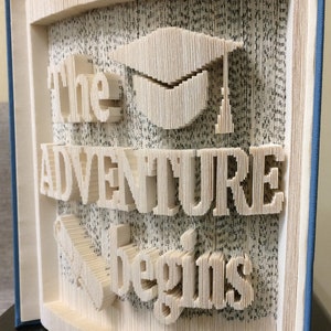 May include: A book art sculpture with the words "The Adventure Begins" and a graduation cap. The sculpture is made from the pages of a book that have been folded and cut to create the design. The book is bound in blue.