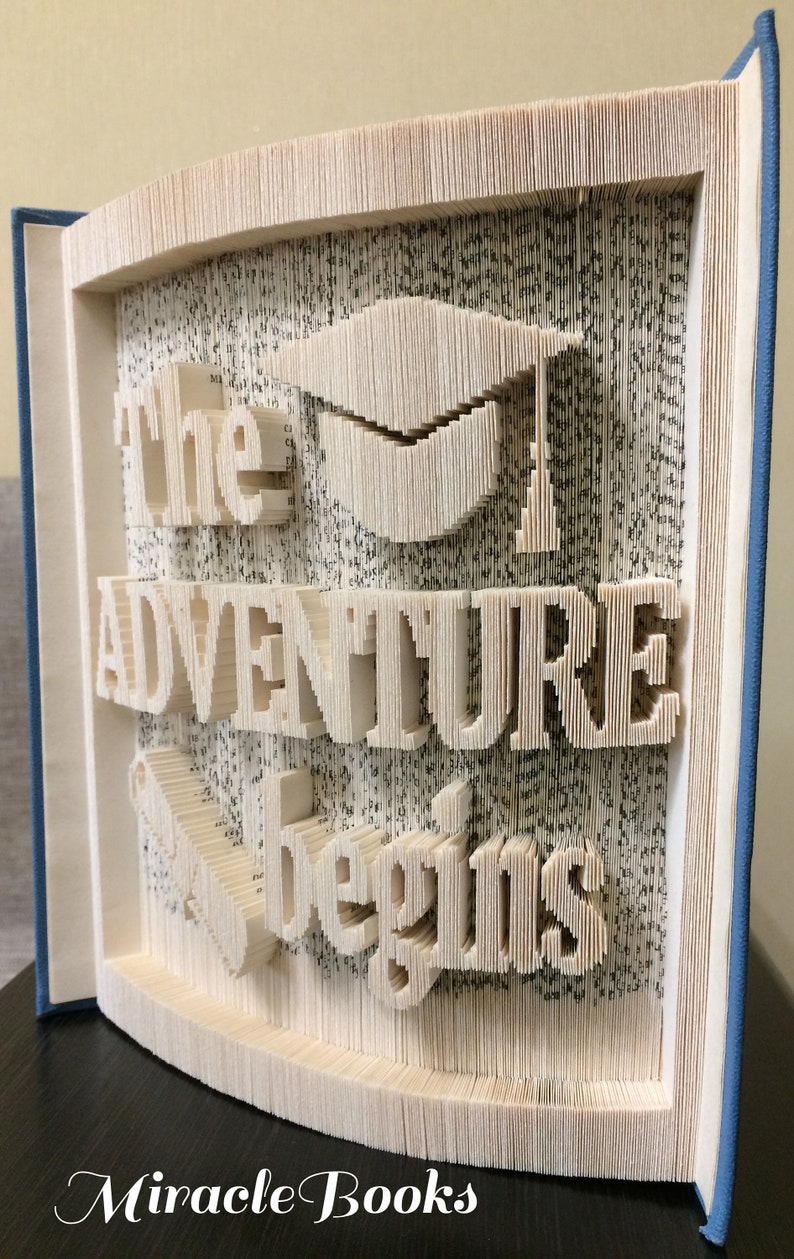May include: A book art sculpture with the words "The Adventure Begins" and a graduation cap. The sculpture is made from the pages of a book that have been folded and cut to create the design. The book is bound in blue.