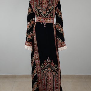 May include: A long black dress with intricate embroidery in shades of pink, red, green, and gold. The dress has a round neckline and long sleeves with a floral pattern. The embroidery features geometric designs and floral motifs.