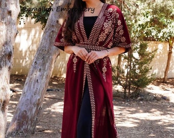 May include: A long, burgundy-colored robe with gold embroidery. The robe has a deep V-neckline and is open in the front. The robe is worn over black leggings and black shoes. The text "Ramallah Embroidery" is visible on the robe.