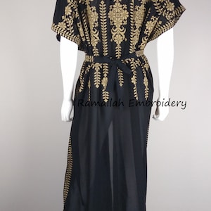 May include: Black kaftan dress with gold embroidery. The dress features intricate floral and leaf patterns on the bodice, sleeves, and along the sides. A matching tie belt cinches the waist. The text "Ramallah Embroidery" is visible.