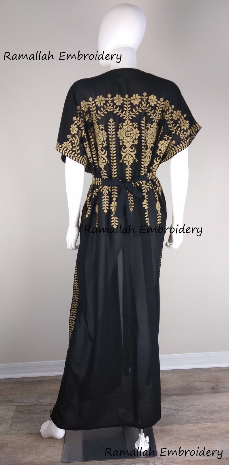 May include: Black kaftan dress with gold embroidery. The dress features intricate floral and leaf patterns on the bodice, sleeves, and along the sides. A matching tie belt cinches the waist. The text "Ramallah Embroidery" is visible.