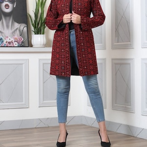 May include: A long, open-front red coat with intricate black embroidery. The coat features a detailed pattern and long sleeves. The person is wearing light blue jeans and black heels. The coat is a statement piece.