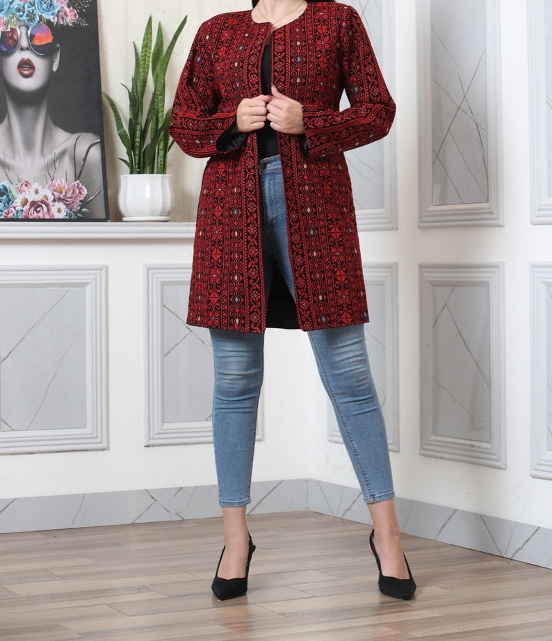 May include: A long, open-front red coat with intricate black embroidery. The coat features a detailed pattern and long sleeves. The person is wearing light blue jeans and black heels. The coat is a statement piece.