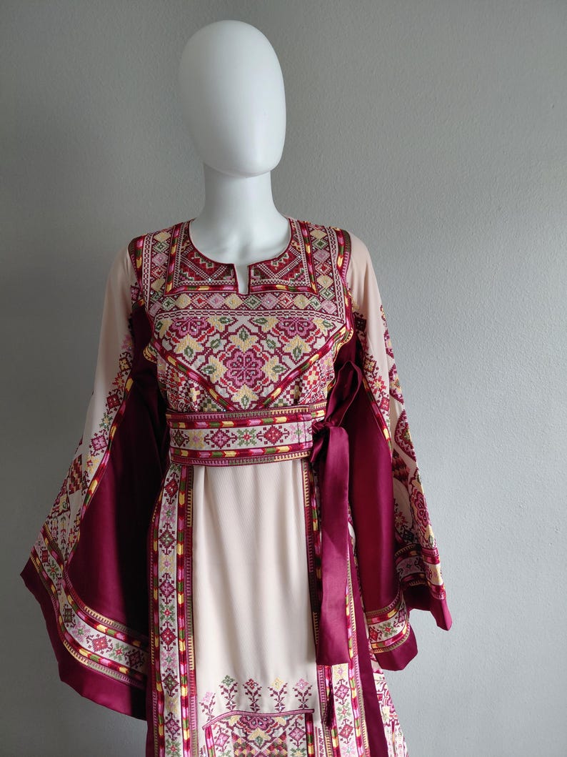 May include: A long-sleeved dress with intricate embroidery in shades of burgundy, pink, and yellow. The dress features a keyhole neckline, a cinched waist with a matching sash, and decorative patterns along the sleeves and hem. The dress is displayed on a mannequin.