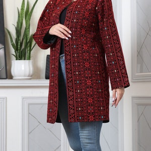 May include: A long, open-front jacket in a vibrant red and black pattern. The jacket features intricate embroidery with floral and geometric designs, and long sleeves. The jacket is worn over a black top and blue jeans.