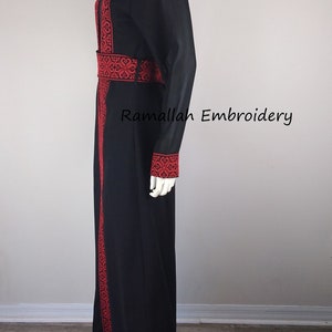 Puede incluir: Una abaya negra con bordados rojos alrededor del cuello, las mangas y la cintura. La abaya est&aacute; en un maniqu&iacute; blanco.