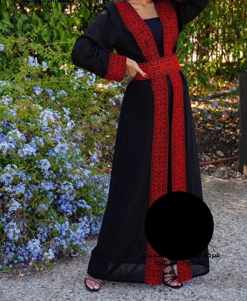 Puede incluir: Una abaya negra con bordados rojos y un cintur&oacute;n rojo. La abaya es larga y fluida, y el bordado es intrincado y detallado. La abaya la lleva puesta una persona de pie delante de un arbusto de flores azules.
